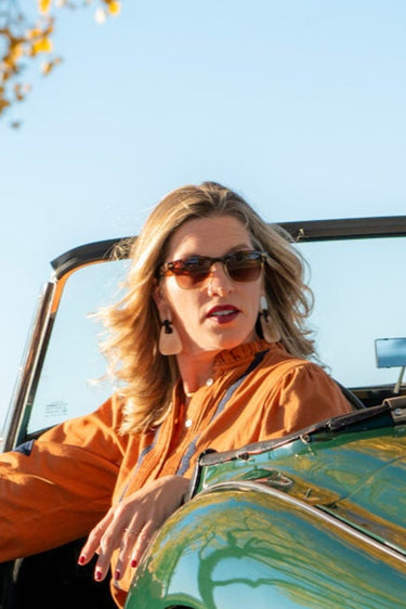 Woman sitting in a green convertible car on a sunny day with trees and a fence in the background.